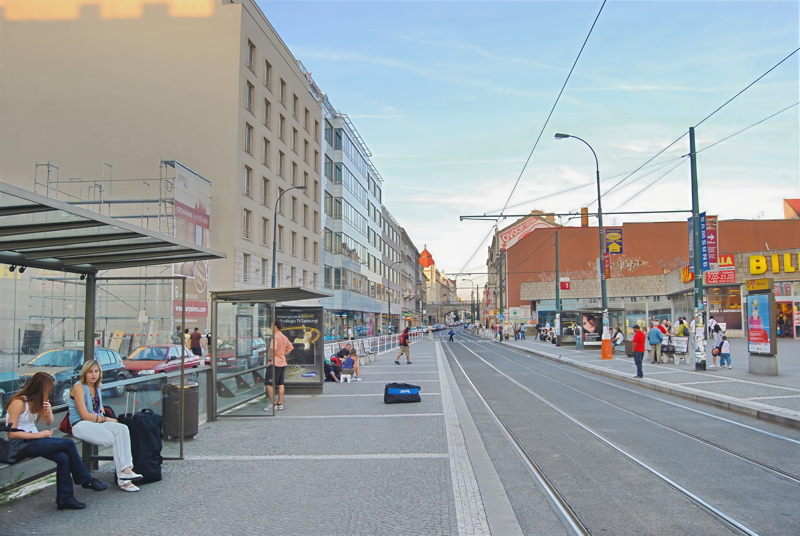 An urban street scene with a tram line, a bus stop shelter, and pedestrians along a wide cobblestone and paved sidewalk
